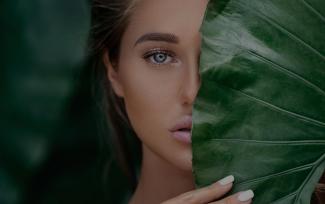Woman with green-blue eyes covers her half face with a large leaf